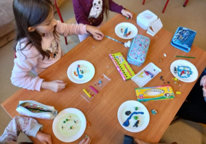 Na zajęciach kreatywnych uczniowie tworzyli jesienny las za pomocą plasteliny. Dzieci siedzą przy stole i ugniatają plastelinę. Na stole są papierowe talerzyki i kolorowa plastelina.